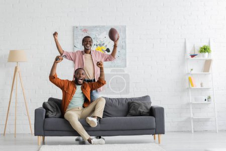 Excited african american father and son watching rugby match in modern living room 
