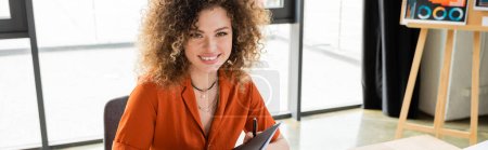 Photo for Cheerful businesswoman with curly hair holding folder in modern office, banner - Royalty Free Image