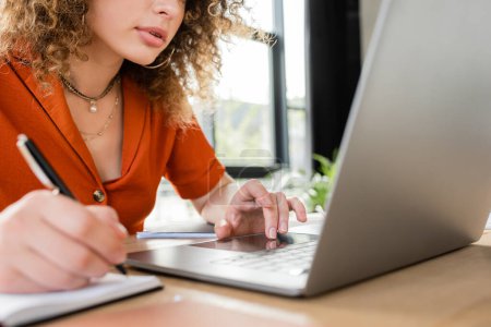 Téléchargez les photos : Vue partielle de la femme d'affaires bouclée prenant des notes dans un carnet tout en utilisant un ordinateur portable - en image libre de droit