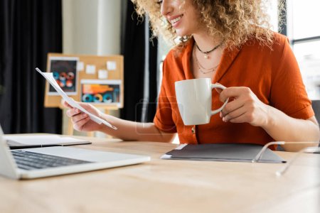 cropped view of cheerful businesswoman with curly hair holding cup of tea near laptop 