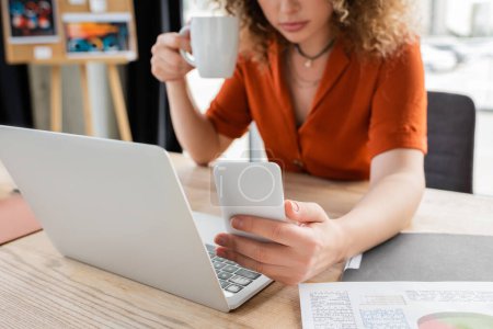 Photo for Cropped view of businesswoman holding cup of tea and using smartphone near laptop on desk - Royalty Free Image