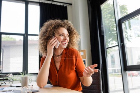 glückliche Geschäftsfrau, die auf dem Smartphone neben verschwommenen Gläsern und einer Tasse Tee auf dem Schreibtisch spricht 