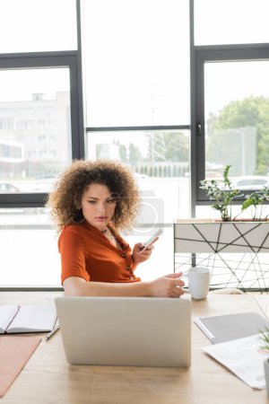 lockige Geschäftsfrau hält Smartphone und Tasse Tee in der Hand, während sie im Büro auf Laptop schaut 