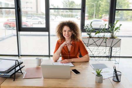 pleased businesswoman with curly hair looking at camera near gadgets and documents on desk 