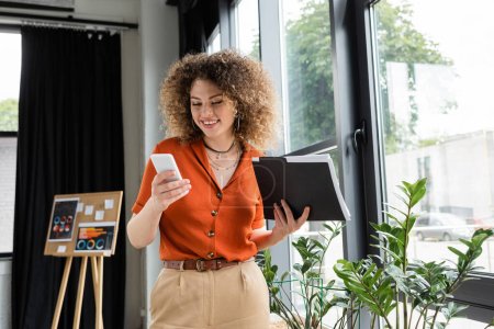 cheerful businesswoman with curly hair holding folder and using smartphone in office 