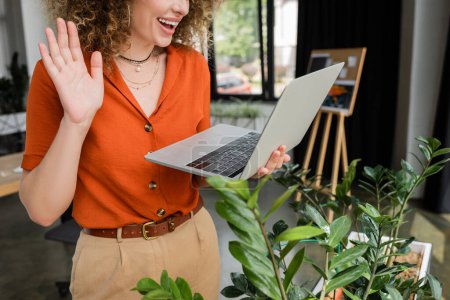 Photo for Cropped view of joyful businesswoman with curly hair having video call and waving hand at laptop in office - Royalty Free Image
