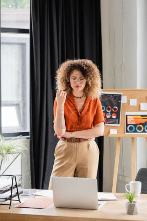 curly businesswoman in glasses looking at laptop near office board with infographics 