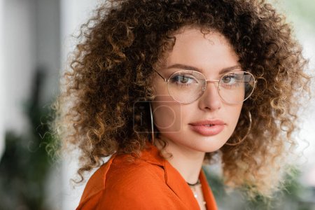 portrait of curly businesswoman in glasses and hoop earrings looking at camera in office 