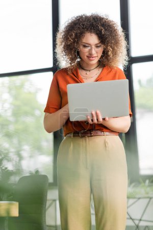 lockige Geschäftsfrau mit Brille lächelt, während sie Laptop im Büro benutzt 