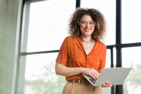 curly businesswoman in glasses smiling while holding laptop in office 