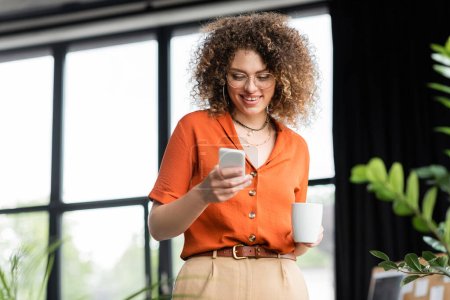 freudige Geschäftsfrau mit Brille, die auf dem Smartphone Nachrichten sendet, während sie im modernen Büro eine Tasse Kaffee hält 