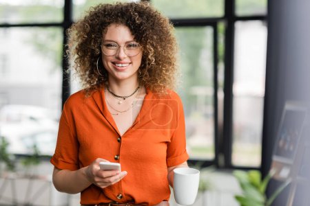 cheerful businesswoman in glasses holding smartphone and cup of coffee while standing in modern office 