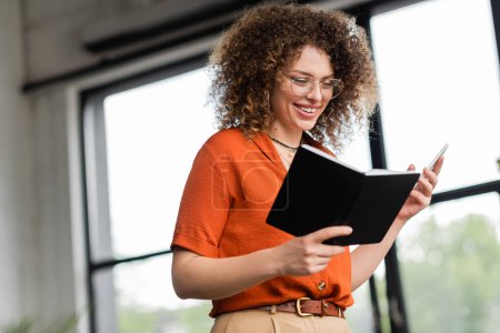 cheerful businesswoman in glasses holding smartphone and notebook while standing in modern office 