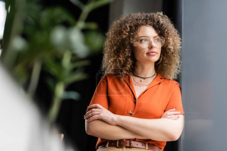 pensive businesswoman with curly hair standing with folded hands in modern office 