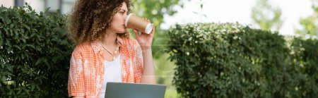 mujer joven con el pelo rizado beber café para ir y el uso de la computadora portátil al aire libre, pancarta 