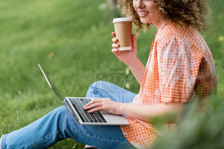 Ausgeschnittene Ansicht einer fröhlichen Freiberuflerin mit lockigem Haar, die Pappbecher in der Hand hält und Laptop benutzt, während sie auf Gras sitzt 