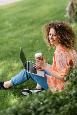 Freelancer mit lockigem Haar hält Pappbecher in der Hand und bedient Laptop im Sitzen auf Gras 