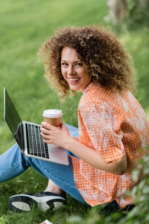 freelancer lleno de alegría con el pelo rizado sosteniendo la taza de papel y el uso de la computadora portátil mientras está sentado en la hierba 