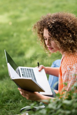 Téléchargez les photos : Femme concentrée avec des notes de lecture de cheveux bouclés tout en utilisant un ordinateur portable dans le parc - en image libre de droit