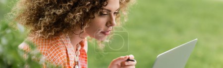 Photo for Focused woman with curly hair using laptop and holding pen in park, banner - Royalty Free Image