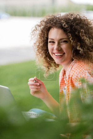 positive freelancer woman with curly hair using laptop and holding pen in park