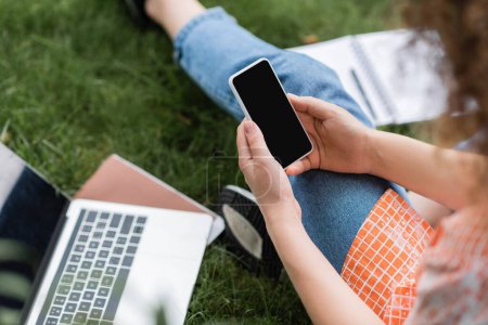 cropped view of young woman holding smartphone with blank screen while sitting near laptop and notebook on grass 