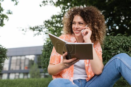 heureuse jeune femme avec bouclé tenant carnet tout en parlant sur smartphone dans le parc vert 