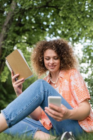 low angle view of smiling young woman with curly holding notebook while using smartphone in green park 