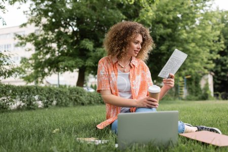femme gaie avec les cheveux bouclés lecture journal et tenant le café pour aller tout en étant assis sur l'herbe près de l'ordinateur portable 