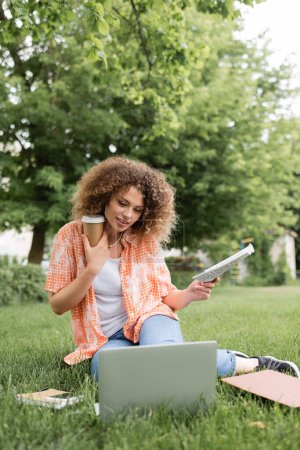 fröhliche Frau mit lockigem Haar, die Zeitung und Kaffee to go in der Hand hält, während sie auf dem Gras sitzt und auf Laptop schaut 