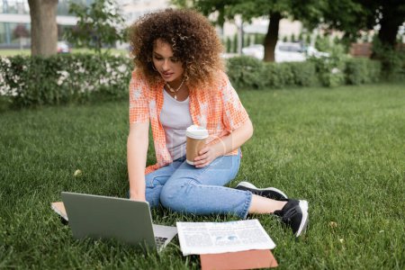 Téléchargez les photos : Jeune femme avec les cheveux bouclés tenant café pour aller tout en étant assis sur l'herbe et en utilisant un ordinateur portable - en image libre de droit