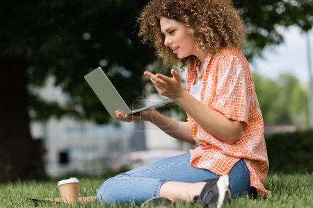 young cheerful woman with curly hair holding laptop while working remotely in green park 