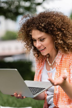 Freelancer Frau mit lockigem Haar hält Laptop in der Hand, während sie ferngesteuert im grünen Park arbeitet 