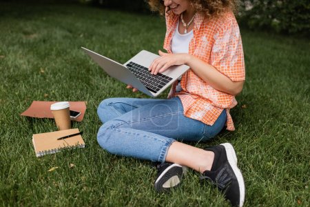 cropped view of happy woman using laptop while sitting on lawn near paper cup and notebook 