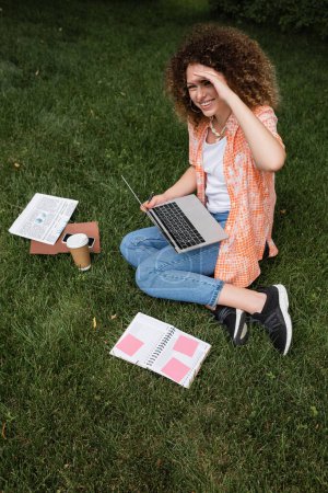 Téléchargez les photos : Vue grand angle de femme freelance joyeux tenant ordinateur portable tout en étant assis sur la pelouse dans le parc - en image libre de droit
