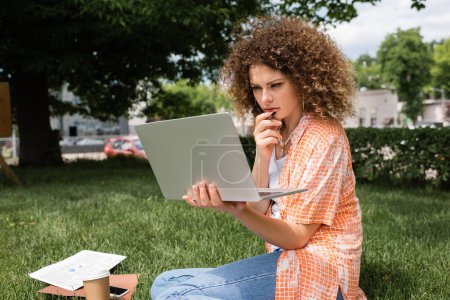 Photo for Pensive freelancer with curly hair holding laptop while sitting in green park - Royalty Free Image