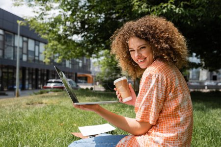 Téléchargez les photos : Joyeux et bouclé pigiste tenant tasse en papier et ordinateur portable tout en travaillant à distance dans le parc - en image libre de droit