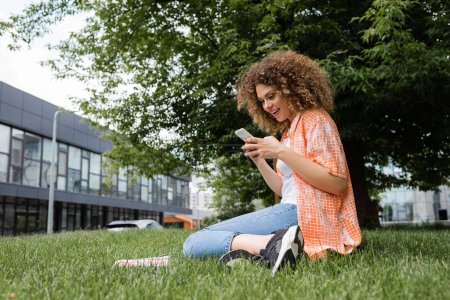 cheerful woman with curly hair using smartphone while sitting on lawn in green park 
