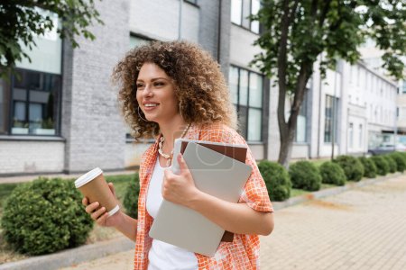 Téléchargez les photos : Femme heureuse avec les cheveux bouclés tenant café pour aller et ordinateur portable avec dossier tout en marchant à l'extérieur - en image libre de droit
