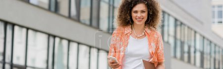 cheerful woman with curly hair holding smartphone and folder while walking outside, banner 