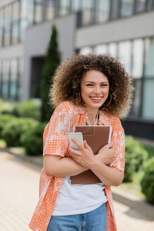 cheerful woman with curly hair holding smartphone and folder while smiling outside 