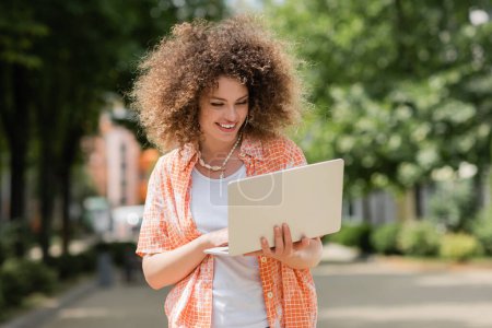 lockige freiberufliche Frau lächelt, während sie Laptop in der Hand hält und ferngesteuert im Park arbeitet 