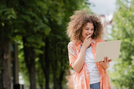 Foto de Feliz freelancer mujer sonriendo mientras sostiene el ordenador portátil y trabajando remotamente en el parque - Imagen libre de derechos