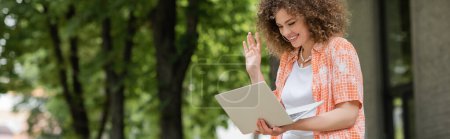 Photo for Cheerful woman smiling while waving hand during video call on laptop in green park, banner - Royalty Free Image