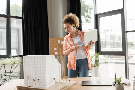 curly architectural designer holding laptop while looking at house model in office 
