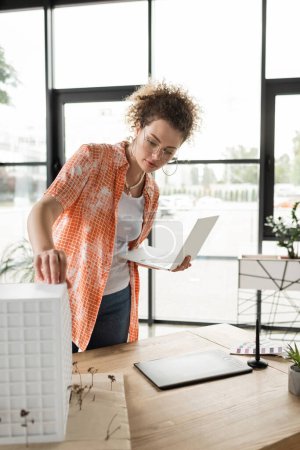 lockige Architekturdesignerin hält Laptop in der Hand, während sie Hausmodell im Büro überprüft 