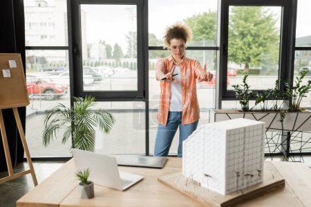 Téléchargez les photos : Jeune designer faisant geste de cadre avec les mains tout en regardant le modèle de maison résidentielle sur le bureau - en image libre de droit