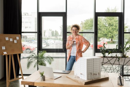 happy architectural designer standing with hand on hip near gadgets and residential house model on desk