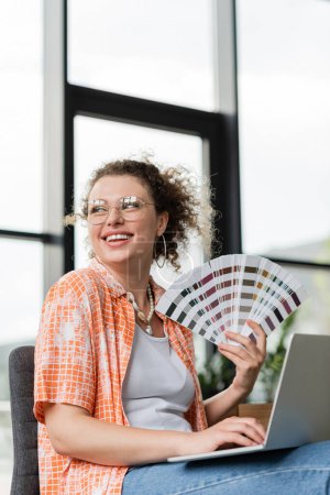happy architectural designer in glasses holding color palette near laptop in office