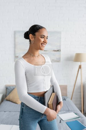 Téléchargez les photos : Étudiant afro-américain souriant tenant ordinateur portable et réserver dans un appartement moderne - en image libre de droit